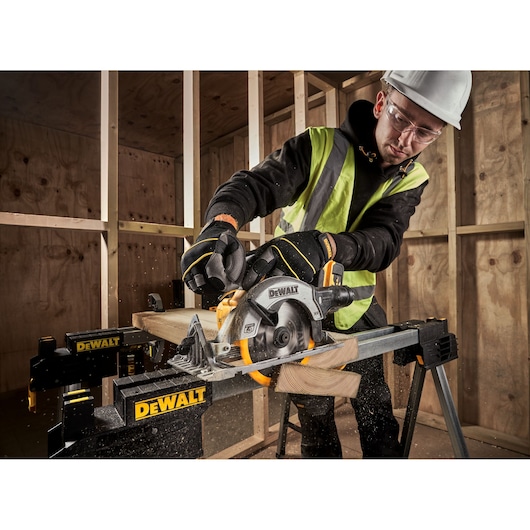 A person wearing safety gear uses a DEWALT cordless circular saw (DCS565P2) to cut wood on a DEWALT workbench in a