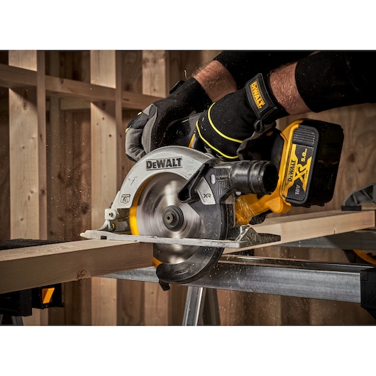 A DEWALT cordless circular saw is being used to cut a wooden plank, with sawdust flying. The tool is held by a person