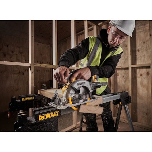 A person wearing a safety vest and helmet operates a DEWALT cordless circular saw to cut a piece of wood on a workbench in a