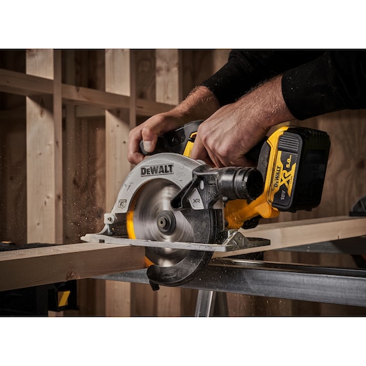 A person using a DEWALT cordless circular saw to cut a wooden plank inside a construction site, with sawdust flying.