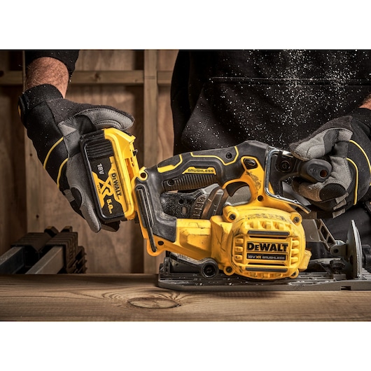Person wearing gloves inserts battery into a yellow DEWALT cordless circular saw on a wooden table.