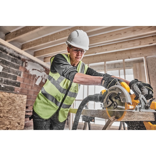 A person wearing a safety vest, gloves, and a hard hat operates a DEWALT DCS570H2T circular saw, cutting wood in a construction site with exposed beams and brick walls.