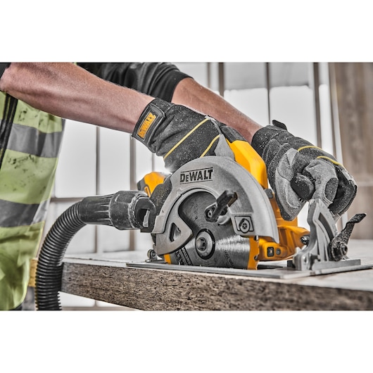 A person wearing work gloves uses a DEWALT circular saw to cut through a piece of wood in a workshop. The saw is connected to a dust extraction hose.