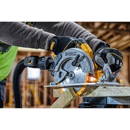 A close-up of a person using a DEWALT DCS570 circular saw to cut a wooden plank, with safety gloves and a dust extraction hose attached, inside a construction site.