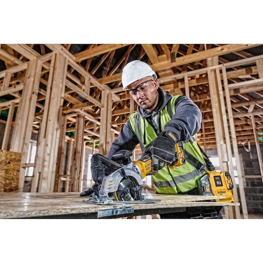 A construction worker wearing a safety helmet and high-visibility vest uses a DEWALT DCS571P2 cordless circular saw to cut wood in a building under construction. The DEWALT tool and battery pack are visible on the workbench.