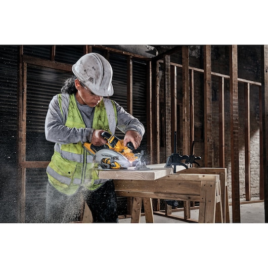 A construction worker wearing a safety helmet and high-visibility vest is using a DEWALT cordless circular saw to cut wooden planks on a workbench in an unfinished building.