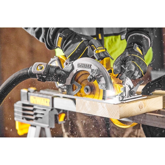 Person uses a DEWALT circular saw to cut wood, wearing safety gloves and connecting to a dust extraction hose.