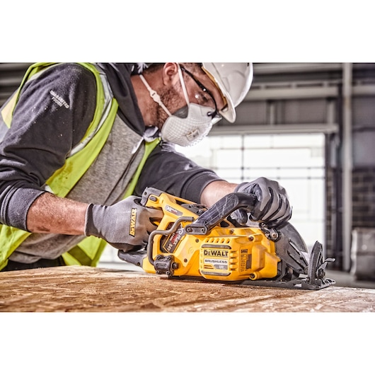 A person wearing safety gear uses a DEWALT DCS577N cordless circular saw to cut a wooden board in an indoor construction setting.