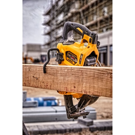 A DEWALT DCS577N cordless circular saw hanging on a wooden beam at a construction site, with scaffolding and building materials visible in the background.
