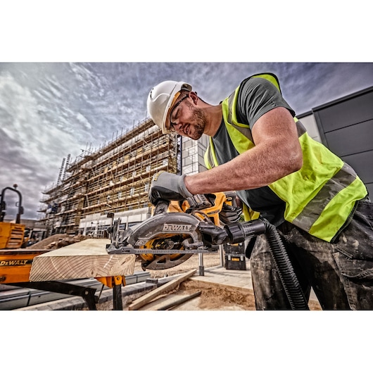 A construction worker wearing a helmet and high-visibility vest is using a DEWALT cordless circular saw (model DCS577T2) to