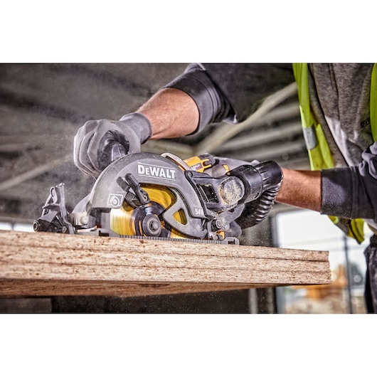 A close-up of a person using a DEWALT DCS577T2 circular saw to cut a piece of wood, with sawdust visible in the air. The