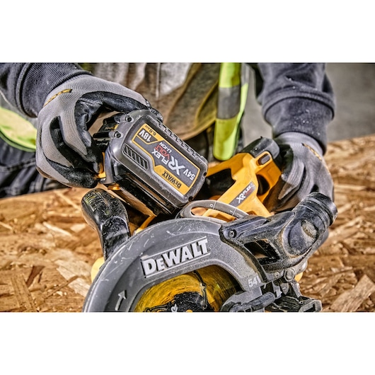 A close-up view of a person wearing gloves and a safety vest, holding a DEWALT cordless circular saw with an 18V XR FLEXVOLT