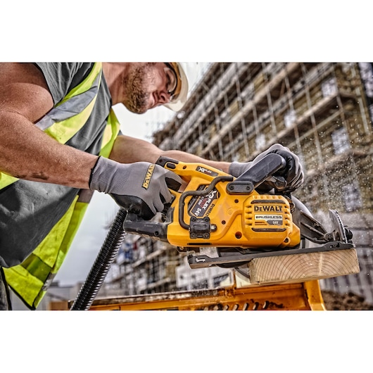 A worker wearing gloves and a high-visibility vest uses a DEWALT cordless brushless circular saw to cut a piece of wood on a