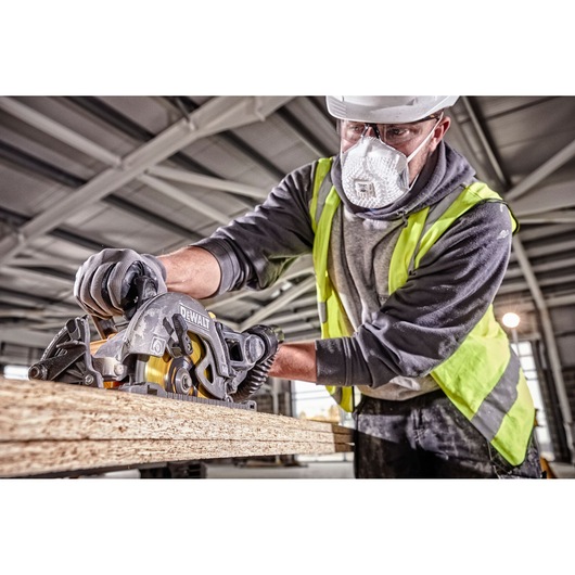 A person wearing work gloves and a high-visibility vest is using a DEWALT DCS577T2 circular saw to cut a piece of wood in an