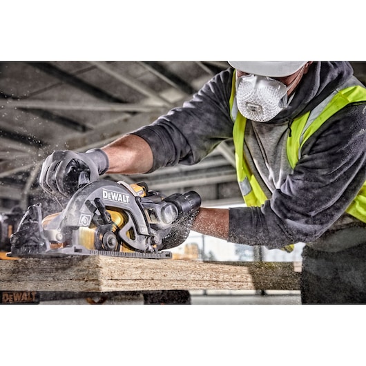 A worker wearing a safety mask, gloves, and a high-visibility vest is using a DEWALT circular saw to cut a wooden plank in