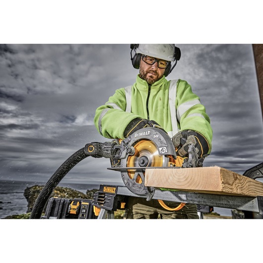 Person using DEWALT DCS578X2 circular saw to cut wood outdoors, wearing safety gear and high-visibility jacket.