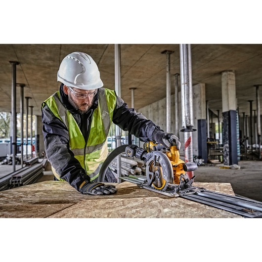 A person wearing a safety helmet, high-visibility vest, and gloves uses a DEWALT DCS579NT circular saw to cut wood on a