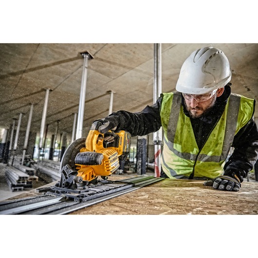 Worker uses DEWALT DCS579N circular saw on wood at a construction site, wearing safety gear.