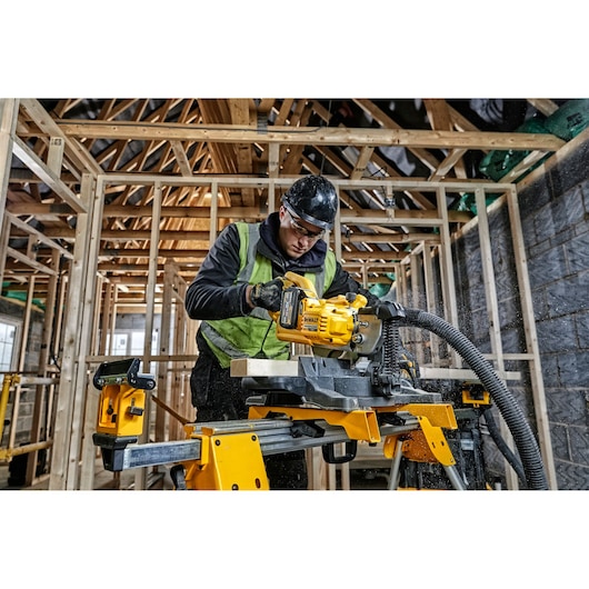 A worker using a DEWALT DCS777T2 electric saw to cut wood inside a house under construction. The worker is wearing safety gear and a green vest. The surrounding area features wooden framing and construction materials.