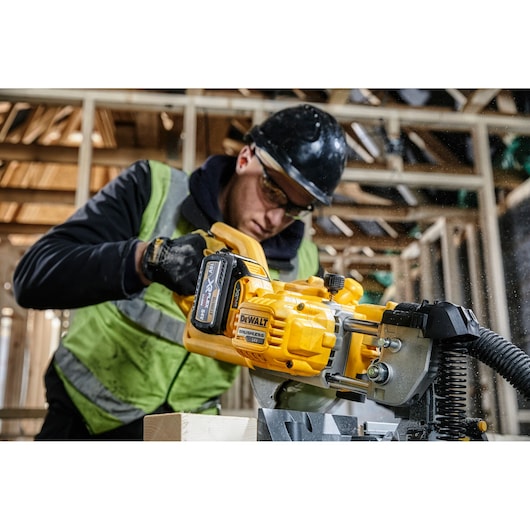 A person wearing safety gear operates a DEWALT DCS777T2 power tool, cutting wood in a construction site. The tool is bright yellow, and the person is in a partially built wooden structure.