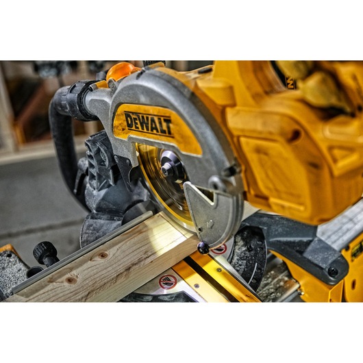 Close-up of a DEWALT miter saw cutting a piece of wood, showing the blade and the wood held in place in a workshop setting.
