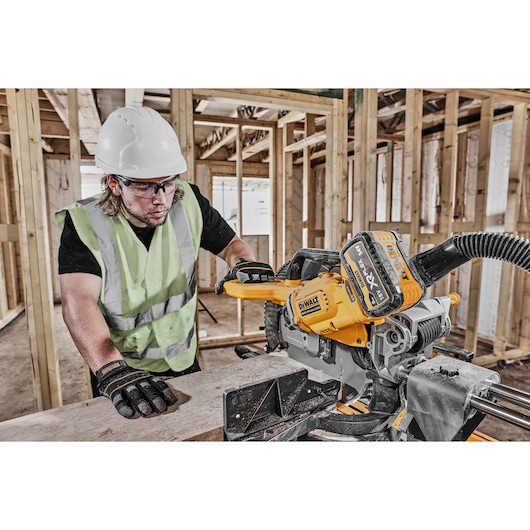 Worker using DEWALT DCS781N saw on a wooden plank at a construction site, wearing safety gear.