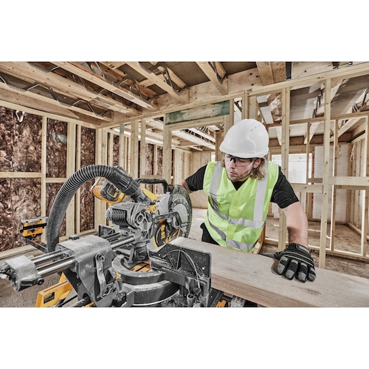 Worker uses DEWALT miter saw DCS781N on wood in a construction site wearing safety gear.