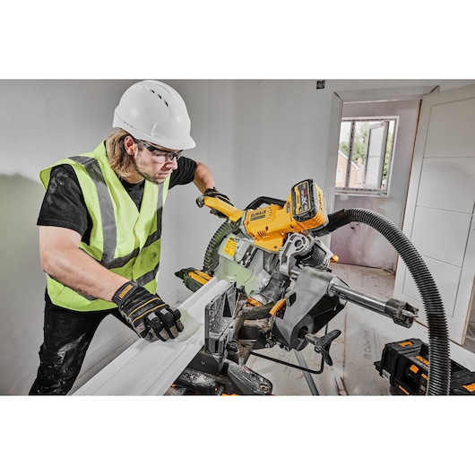 Worker uses DEWALT DCS781N saw to cut wood indoors, wearing safety gear and high-visibility vest.