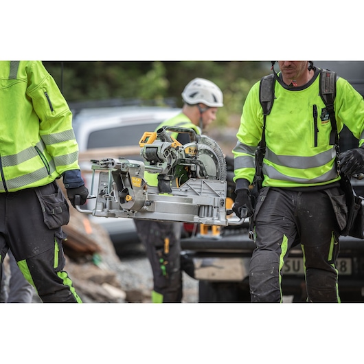 Two workers in safety gear carry a DEWALT miter saw at a construction site.