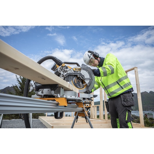 Person using a DEWALT miter saw to cut wood outdoors, wearing safety gear and high-visibility clothing.