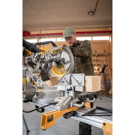 DEWALT miter saw cutting wood in a workshop, operated by a person wearing a green cap and plaid shirt.