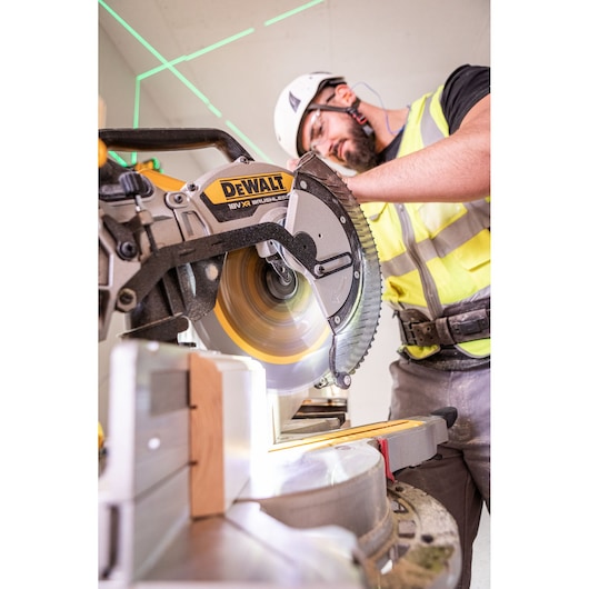 Person using a DEWALT miter saw to cut wood, wearing safety gear at a construction site.