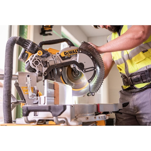 Person using a DEWALT miter saw to cut wood indoors, wearing safety gear and a high-visibility vest.