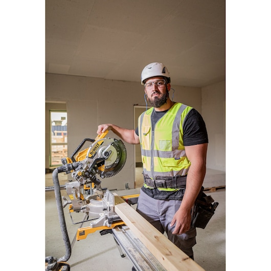 Worker in safety vest using a DEWALT DCS782N mitre saw indoors on a wooden plank at a construction site.