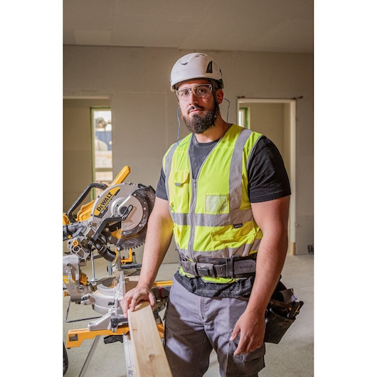 Person wearing safety gear beside a DEWALT DCS782N saw, working with wood inside a construction site.