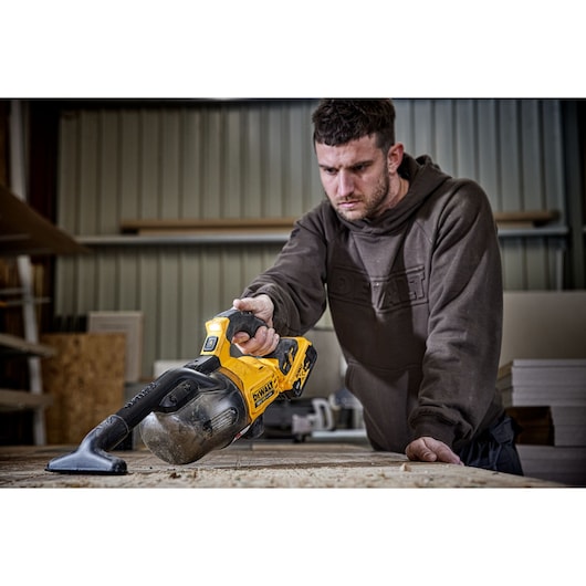 A person using a DEWALT DCV501LN cordless handheld vacuum cleaner to clean a workbench in a workshop environment. The tool is yellow and black, and dust is visible on the work surface.