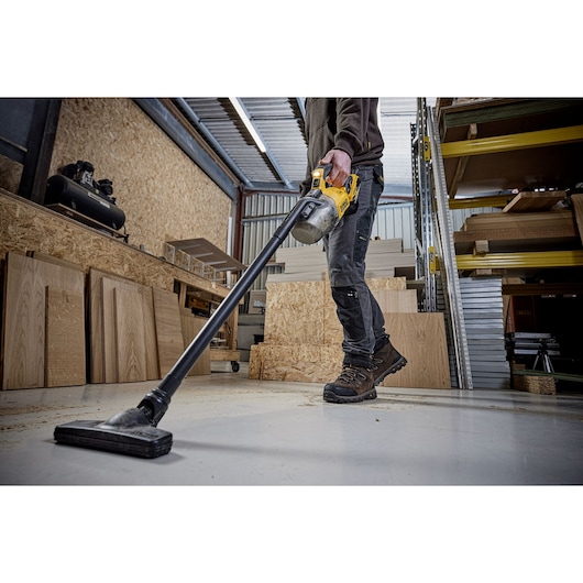 A person using a DEWALT cordless vacuum cleaner (model DCV501LN) to clean the floor of a workshop that has wooden panels and shelves.