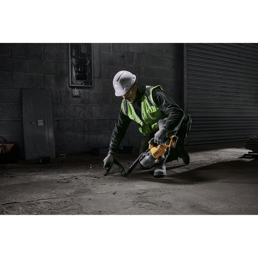 A construction worker wearing a high-visibility vest and hard hat uses a DEWALT portable vacuum cleaner (model DCV501LN) to clean debris from the floor of an industrial site.
