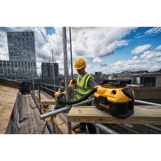 A DEWALT DCV584L portable vacuum cleaner is placed on a wooden platform at a construction site, with a worker wearing a