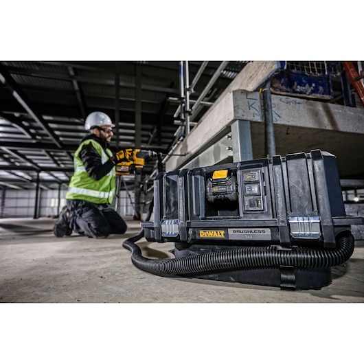 A DEWALT DCV586MT2 54V brushless dust extractor is positioned in the foreground of a construction site, with a worker