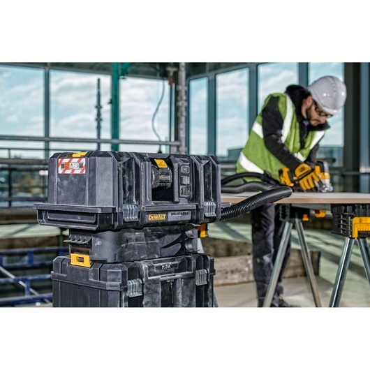 A DEWALT DCV586MT2 dust extractor is shown on a construction site. The unit is connected by hose to a person wearing safety