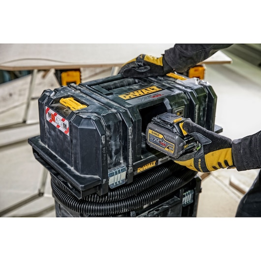 A person wearing work gloves inserts a battery into a DEWALT cordless jobsite dust extractor. The unit is black with yellow