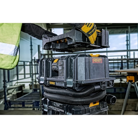 A person wearing gloves and a high-visibility vest is placing a black DEWALT tool case onto a stack of equipment in a