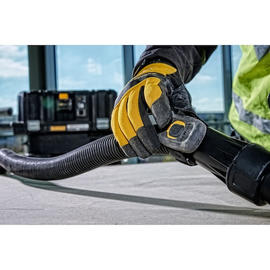Close-up of a person wearing yellow and black work gloves connecting a hose to a DEWALT power tool. A black equipment box is