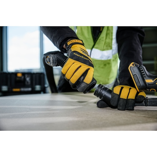 A close-up of a person wearing yellow and black gloves connecting a hose to a DEWALT power tool, with a DEWALT toolbox in