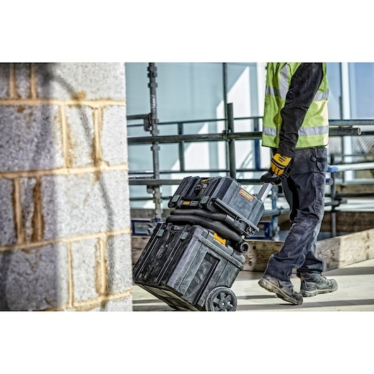 A person wearing work gloves and a high-visibility vest pulls a DEWALT portable vacuum unit with wheels and additional