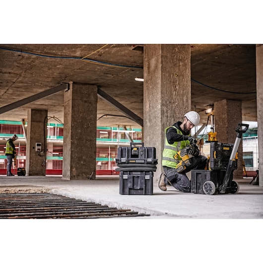 A worker wearing a white helmet and yellow safety vest uses DEWALT tools and storage boxes on a construction site with