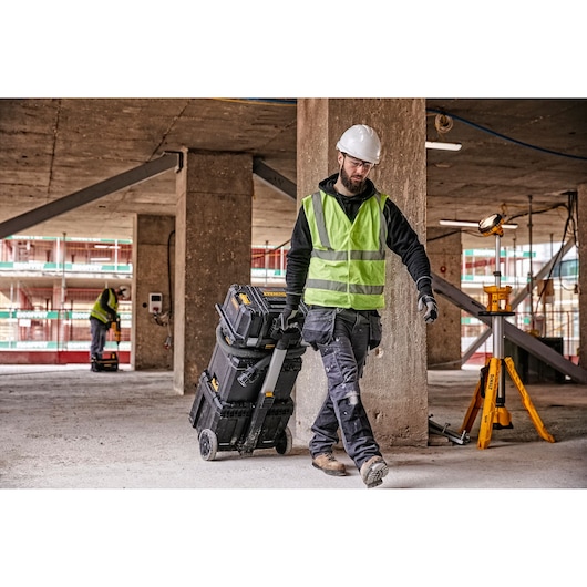 A construction worker wearing a safety helmet and high-visibility vest pulls a DEWALT wheeled toolbox through a building