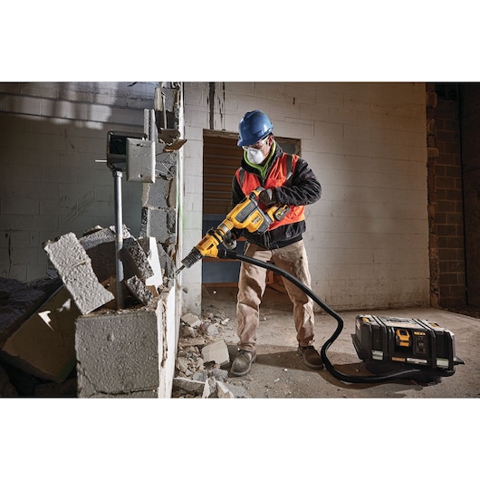 A construction worker wearing a hard hat, safety vest, and face mask uses a DEWALT power tool to break concrete blocks in an