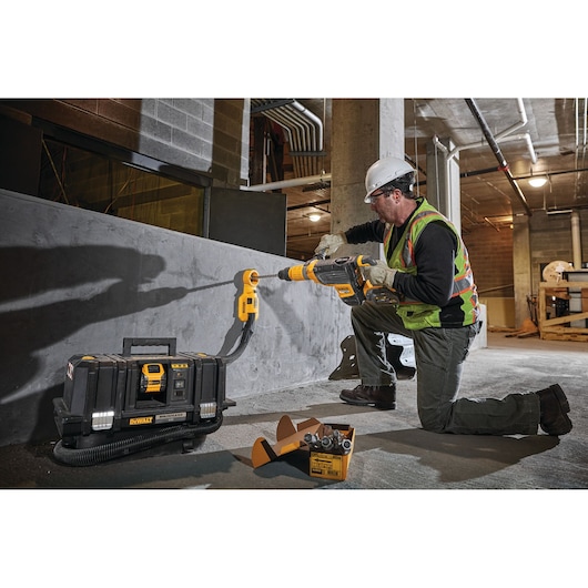 A person wearing a safety vest, gloves, and a hard hat is using a DEWALT cordless vacuum and power tool to drill into a